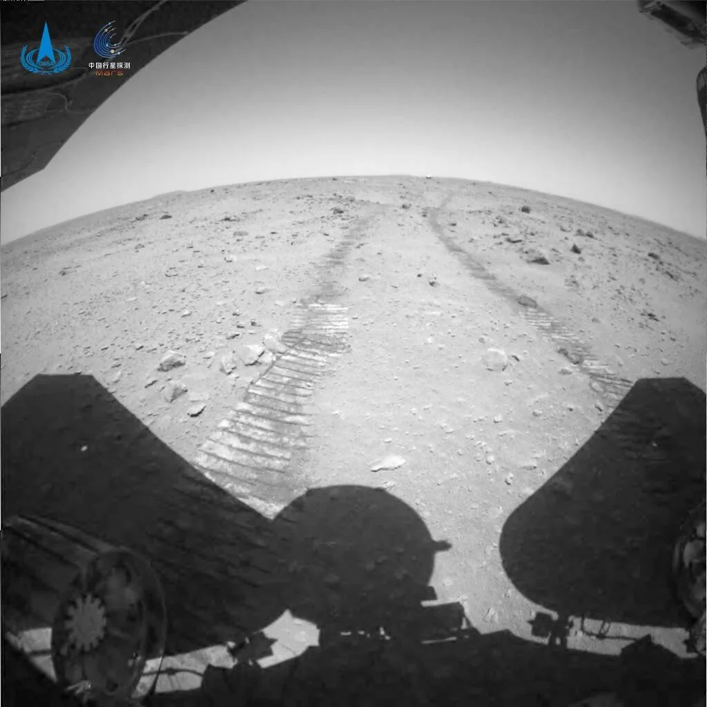 Vista trasera del rover chino mostrando sus huellas sobre el suelo rocoso de Marte, con el logotipo de la misión China Mars en la esquina superior.