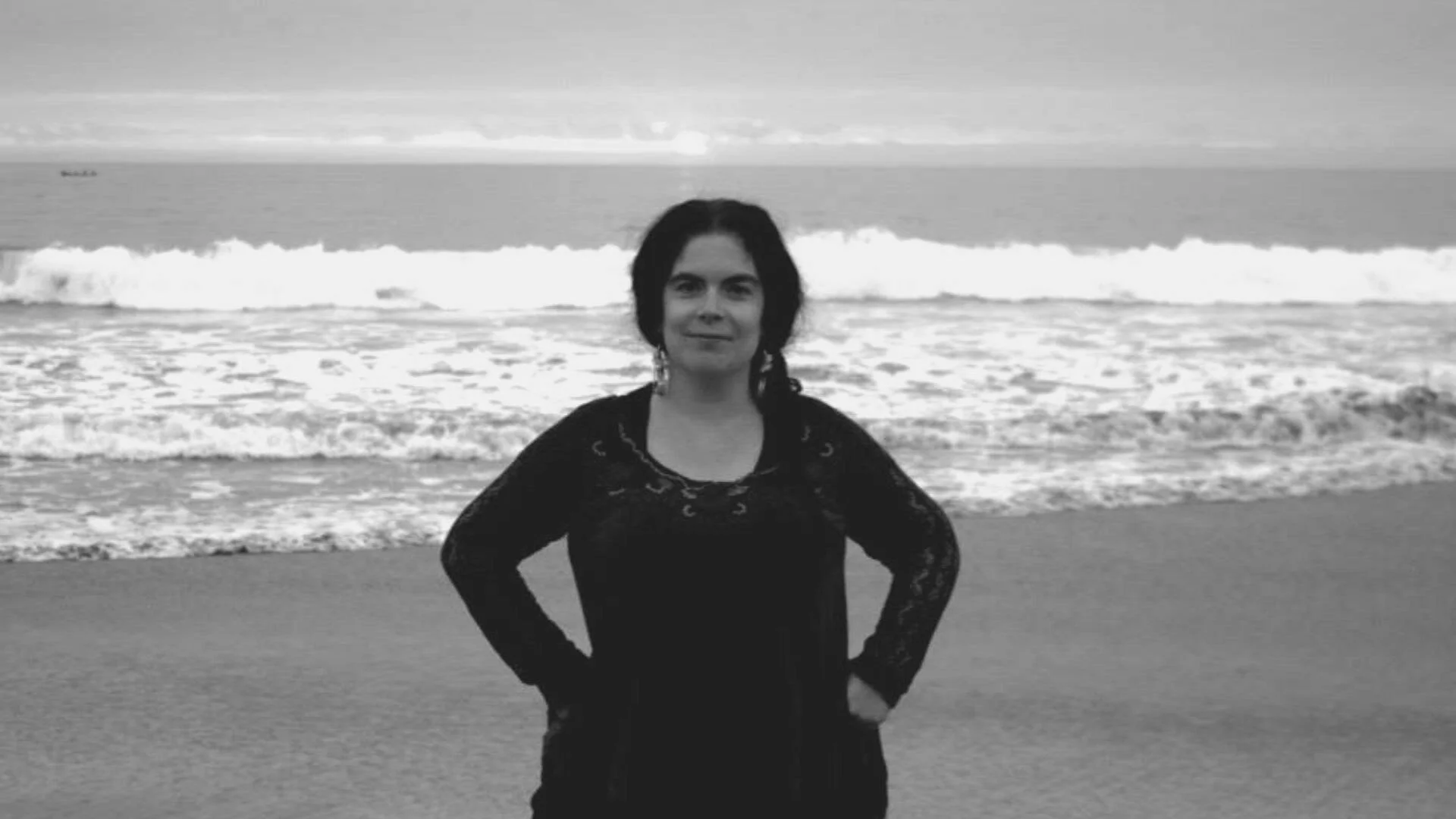 Mujer con vestido negro posa con las manos en la cintura frente a las olas del mar, en fotografía en blanco y negro.