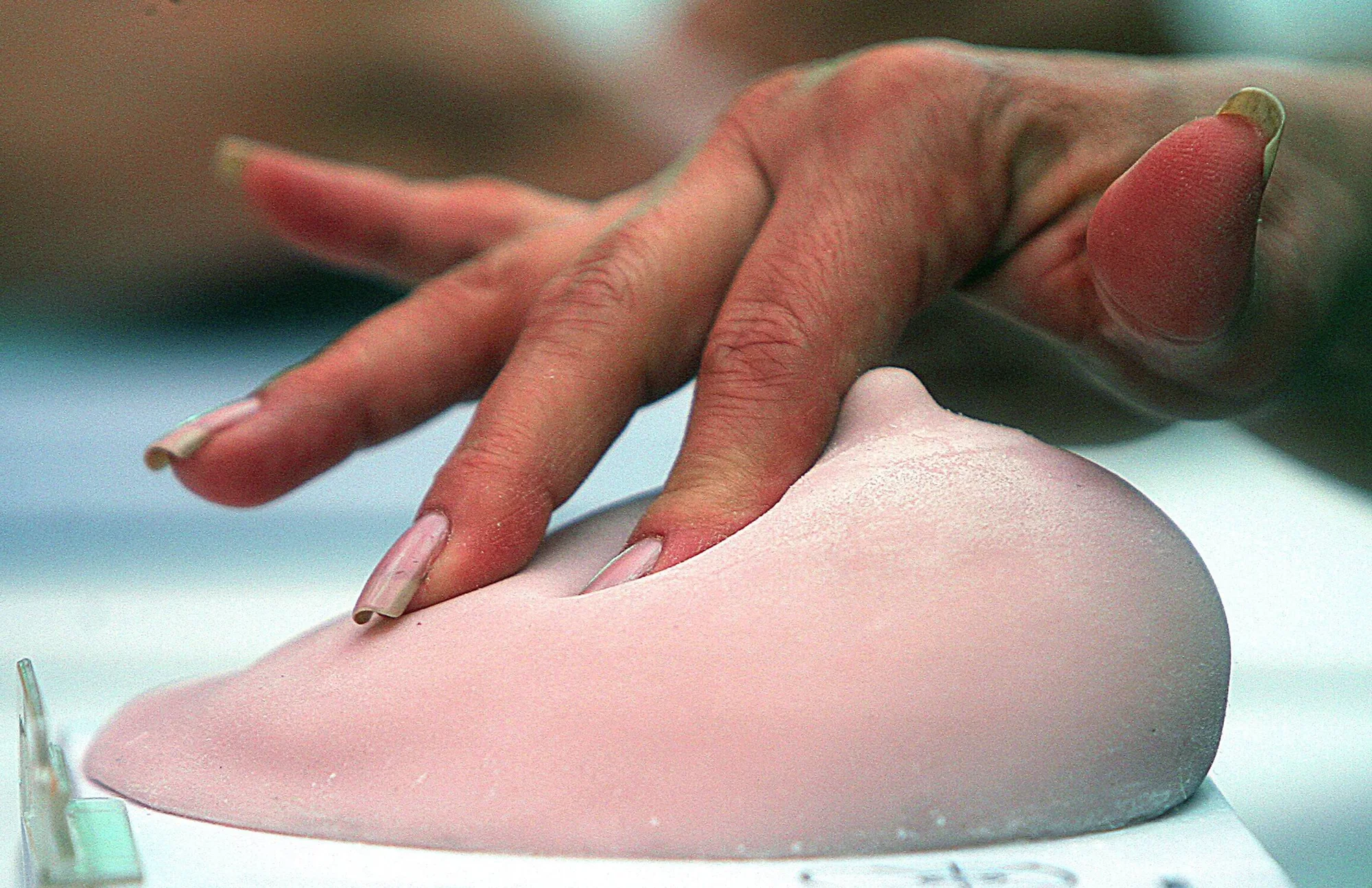 Una mano con uñas largas palpa un implante mamario de silicona rosa sobre una superficie blanca.