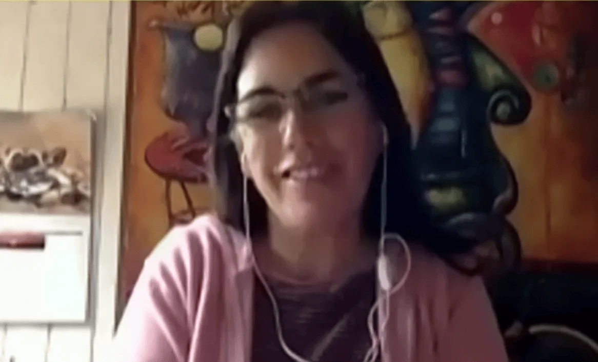Mujer joven con lentes y auriculares blancos sonríe durante una videollamada, con cuadros coloridos al fondo.