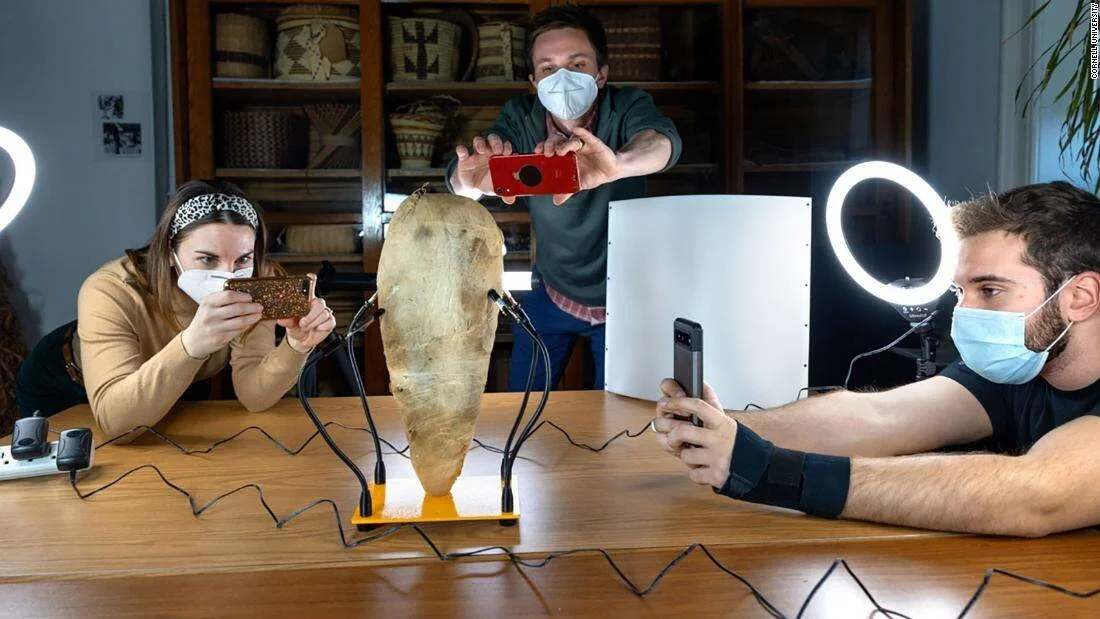 Tres investigadores con mascarilla fotografían con sus móviles una pieza arqueológica de piedra iluminada con focos de anillo sobre una mesa.