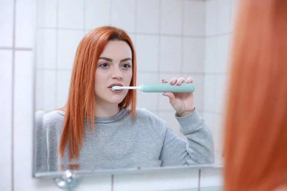 Mujer joven de cabello rojizo se cepilla los dientes con un cepillo eléctrico frente al espejo del baño.