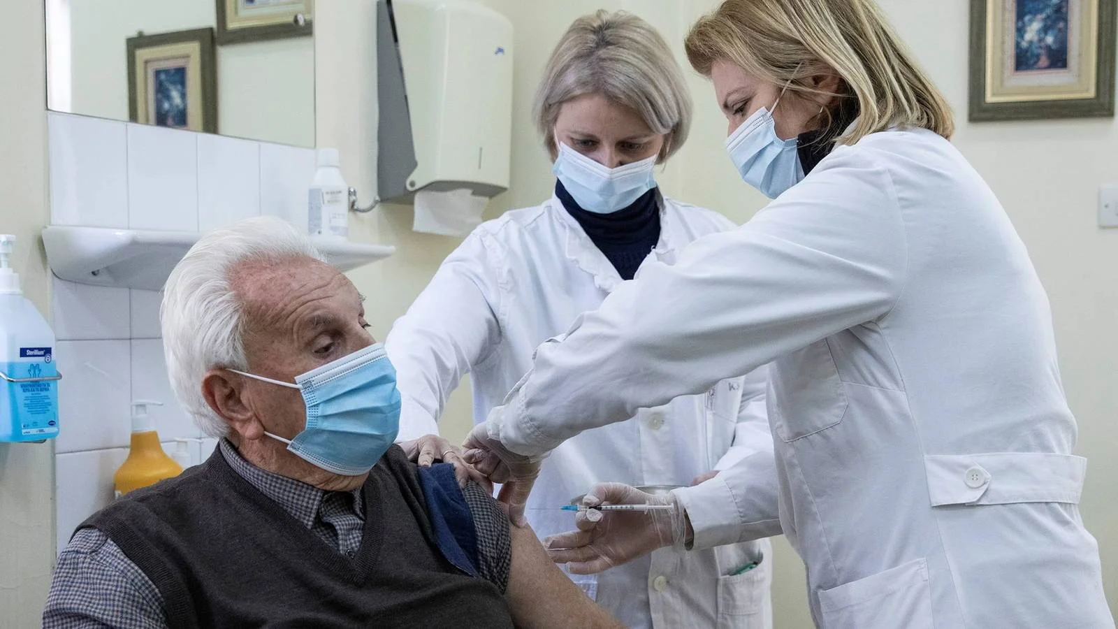 Dos profesionales de salud con mascarilla aplican una vacuna en el brazo a un anciano sentado en un consultorio médico.