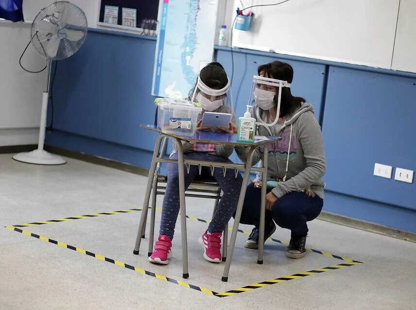 Una maestra con máscara y escudo facial asiste a una niña en un pupitre delimitado con cinta en el piso durante la pandemia.