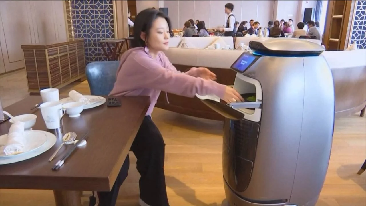 Una mujer retira un plato de comida del compartimento de un robot de servicio autónomo en un restaurante concurrido.