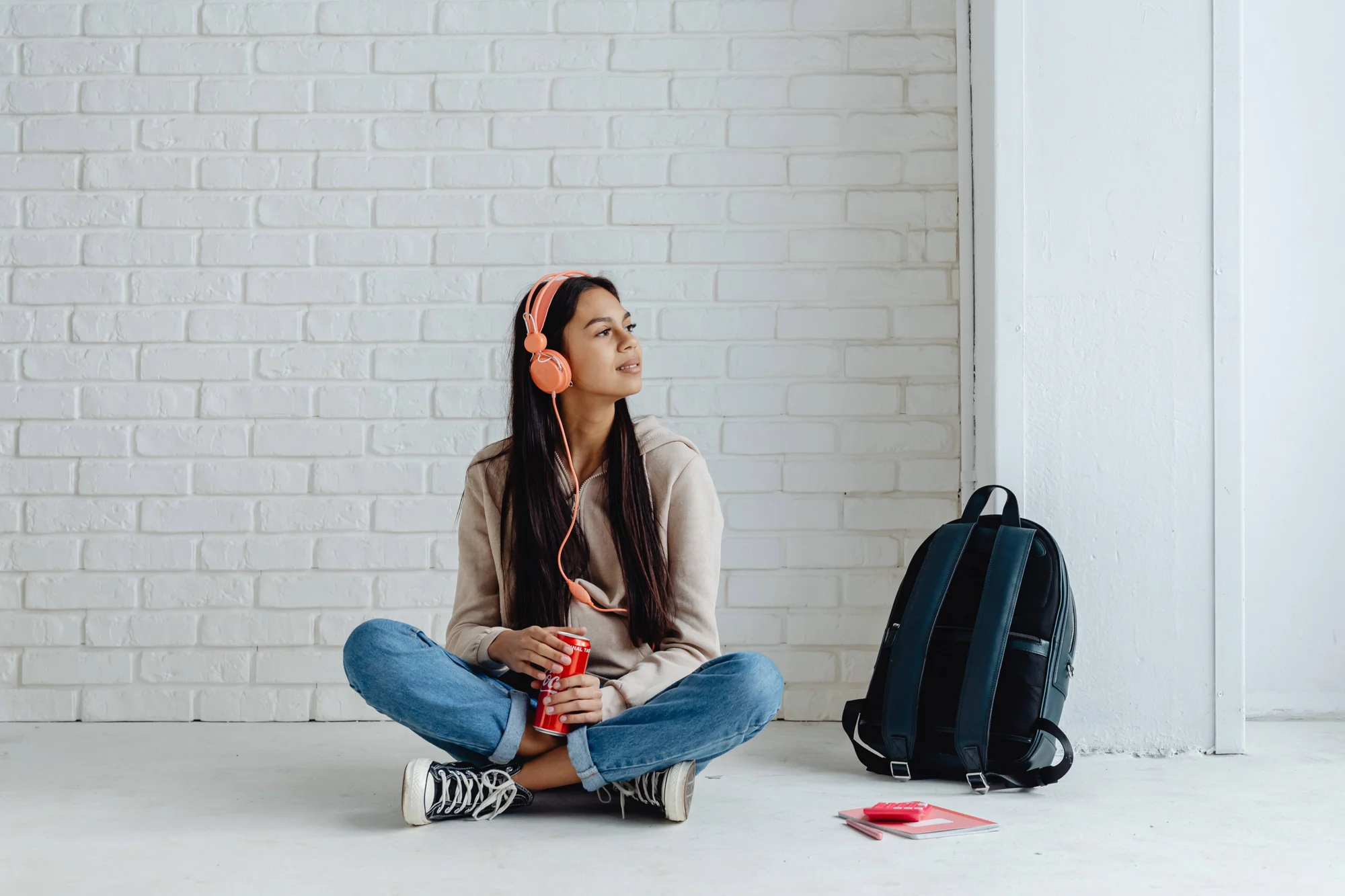 Adolescente sentada en el suelo con auriculares naranjas, sosteniendo una lata de refresco junto a una mochila y cuadernos.