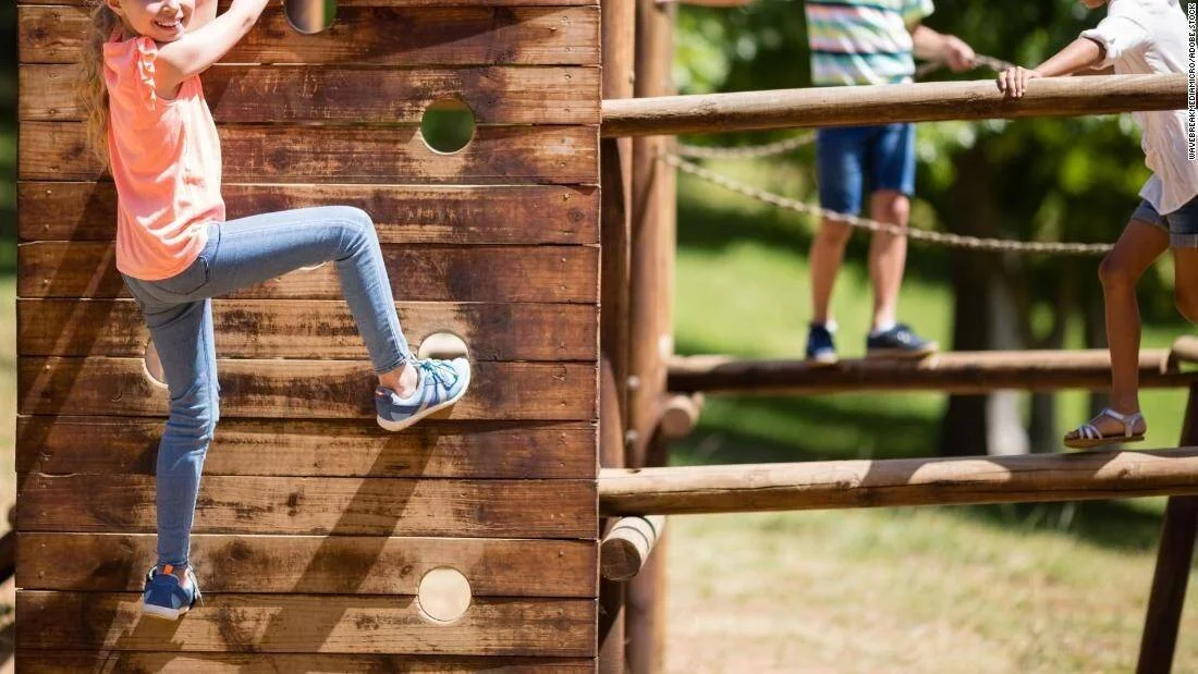 Una niña trepa una pared de madera en un parque infantil mientras otros niños juegan en la estructura.