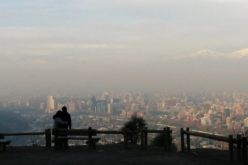 Dos personas sentadas en un banco contemplan una gran ciudad envuelta en smog con montañas nevadas al fondo.