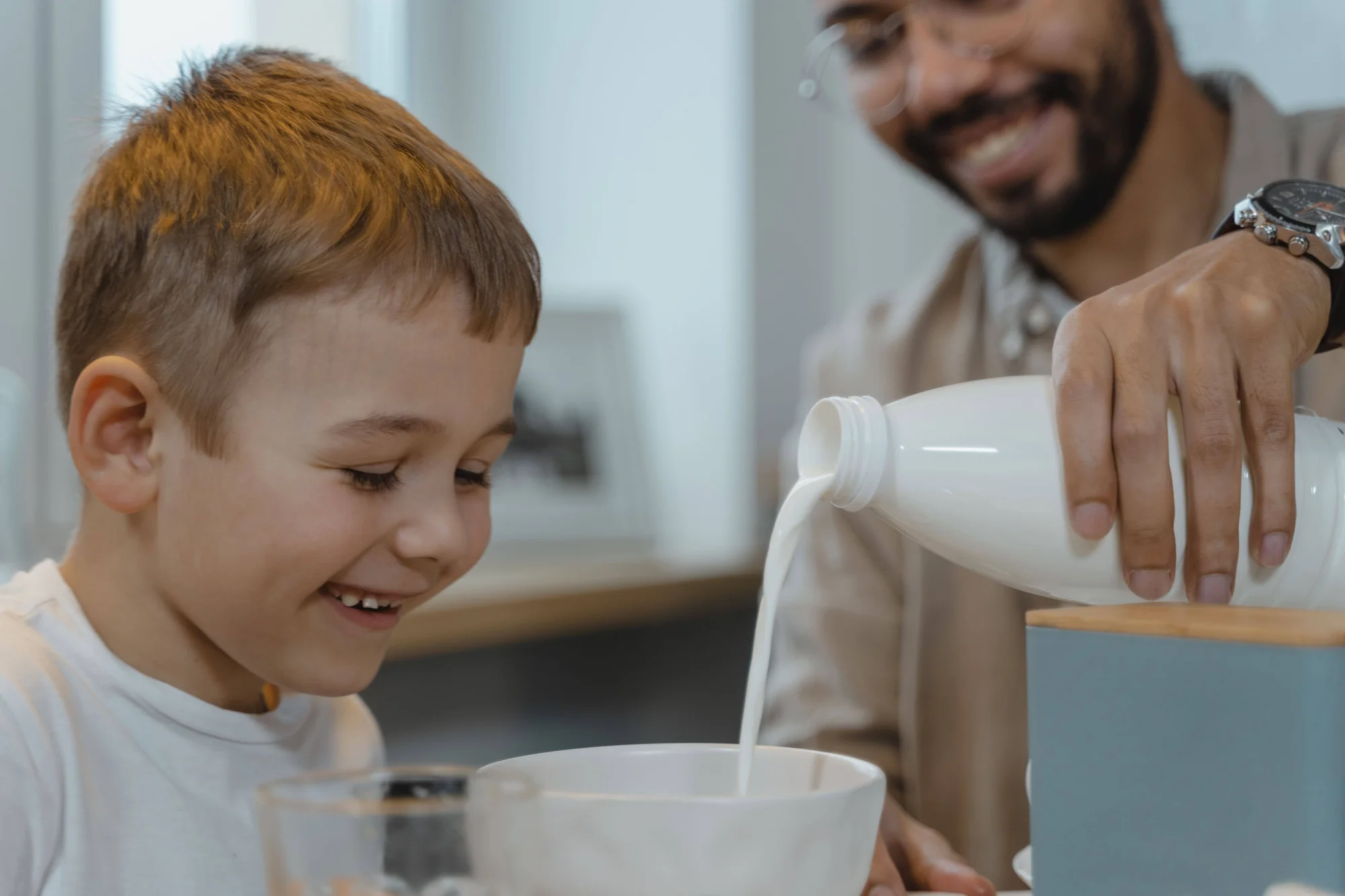 Un adulto vierte leche desde una botella en un tazón blanco mientras un niño pequeño sonríe al verlo.