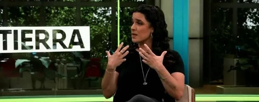 Mujer de cabello oscuro gesticula con las manos durante una entrevista en un estudio de televisión con el rótulo 'Tierra'.