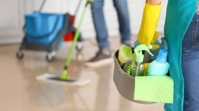 Persona con guantes amarillos sostiene un balde con productos de limpieza mientras otra friega el suelo al fondo.