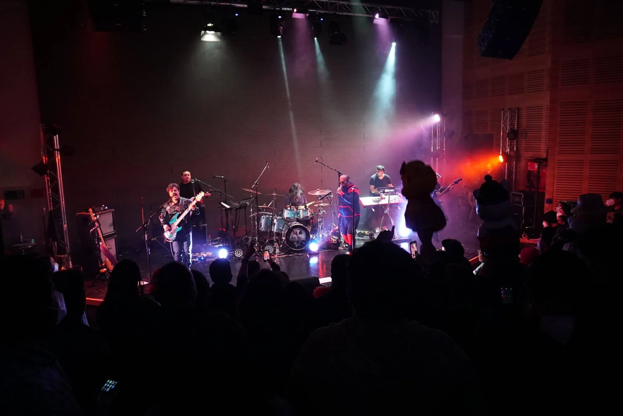 Banda de rock toca en un escenario iluminado con focos de colores ante una multitud de espectadores en sala cerrada.