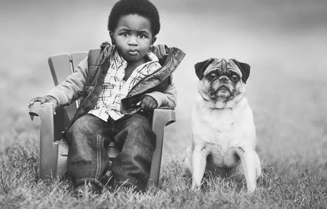 Niño pequeño sentado en una silla al aire libre junto a un perro pug, ambos con expresión seria, en foto en blanco y negro.