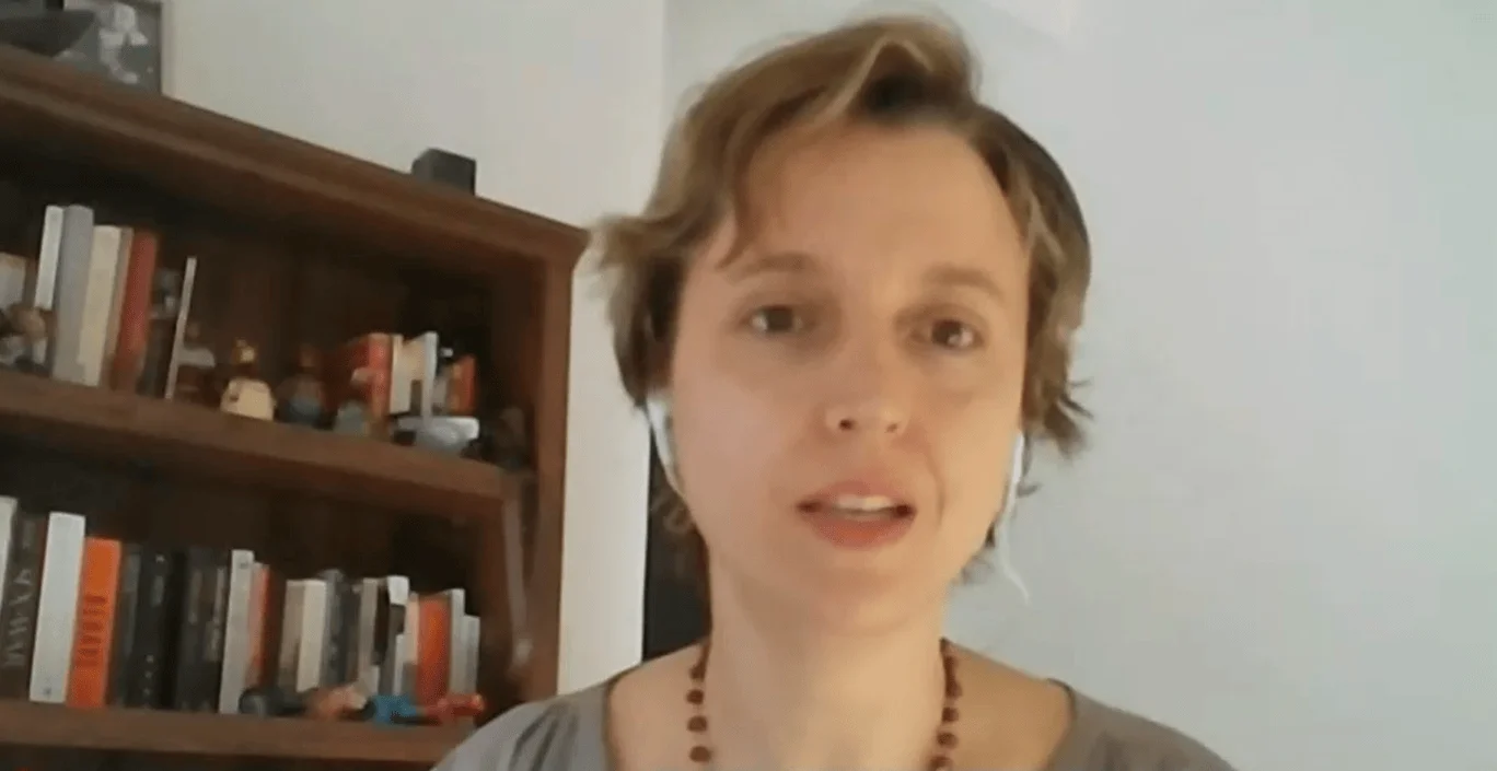Mujer joven con collar de cuentas habla a cámara desde un espacio con estantería llena de libros al fondo.