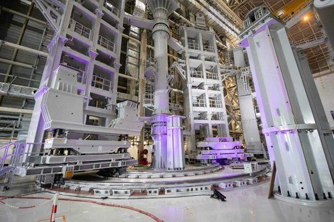 Interior de la obra del reactor de fusión ITER, con grandes estructuras metálicas blancas iluminadas en violeta durante su ensamblaje.