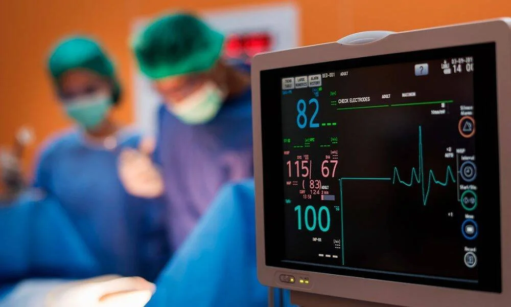 Monitor cardíaco mostrando frecuencia cardíaca y presión arterial mientras cirujanos operan al fondo.
