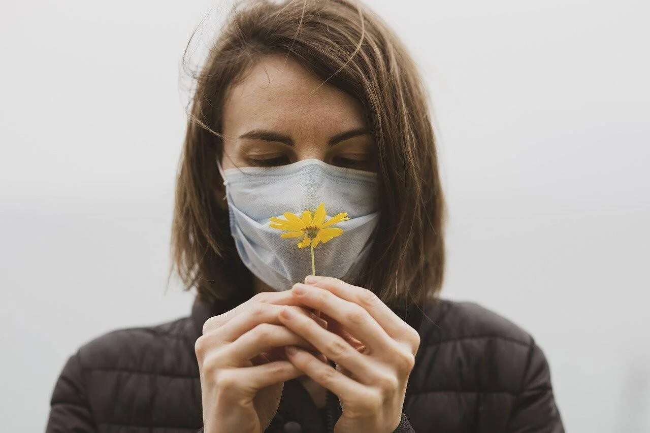 Mujer con mascarilla quirúrgica sostiene una pequeña flor amarilla frente a su rostro, evocando la pérdida del olfato.