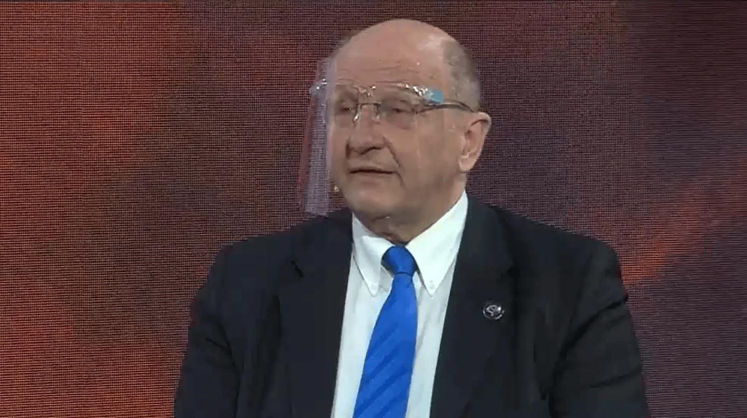 Hombre mayor con anteojos, traje negro y corbata azul habla durante una entrevista en un estudio de televisión.