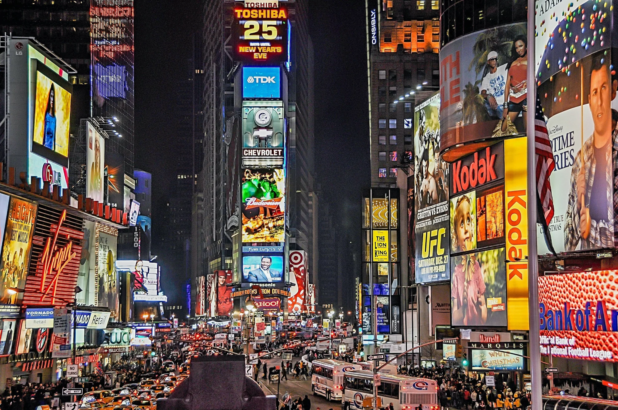 Times Square de Nueva York de noche, con grandes carteles publicitarios iluminados de marcas como Kodak, Chevrolet y Toshiba, y tráfico en la calle.