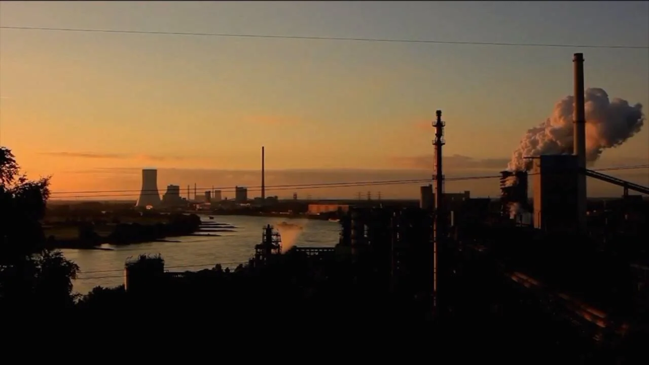Complejo industrial con chimeneas emitiendo humo blanco a orillas de un río bajo un cielo anaranjado al atardecer.
