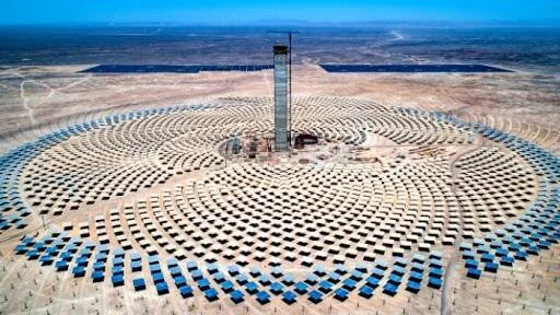 Vista aérea de una planta solar de torre central rodeada de miles de heliostatos dispuestos en forma circular en un desierto.