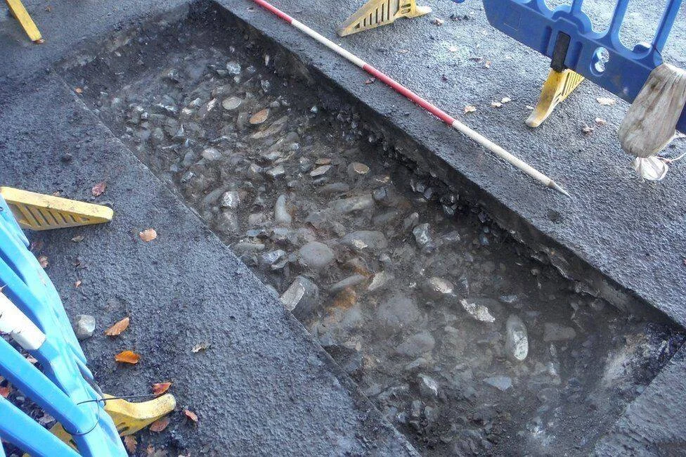 Zanja abierta en una calle asfaltada que revela un antiguo pavimento de piedras bajo el asfalto moderno, con vallas de obra alrededor.