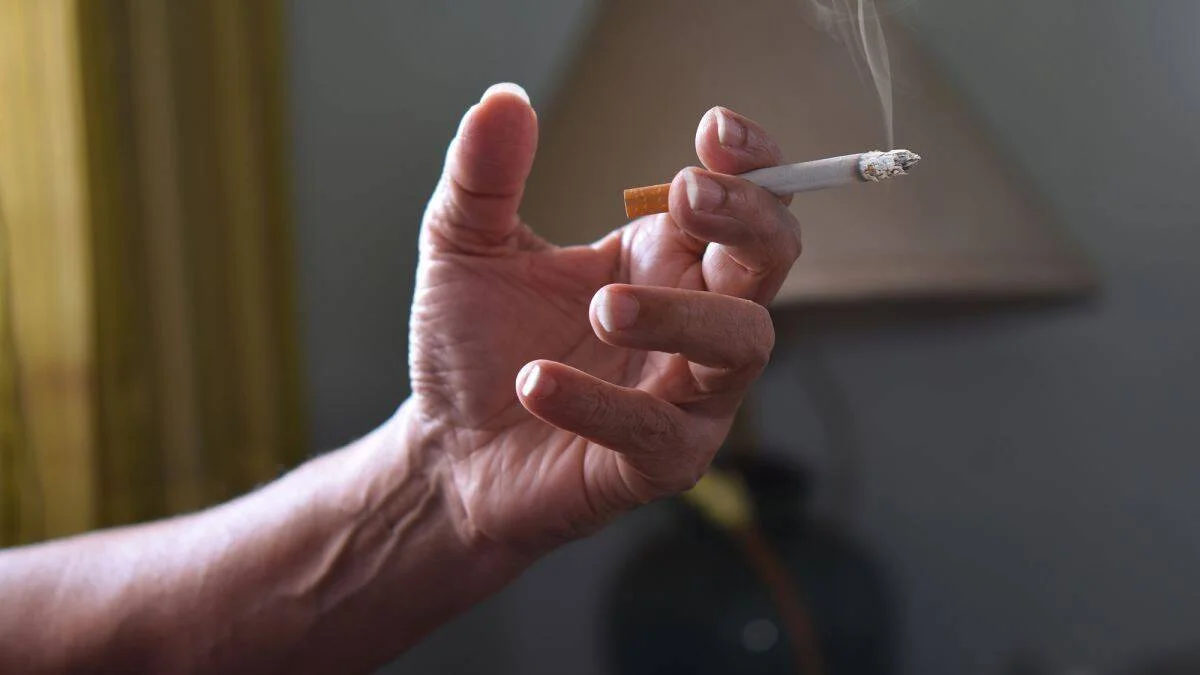 Mano sosteniendo un cigarrillo encendido que desprende humo en un ambiente interior.