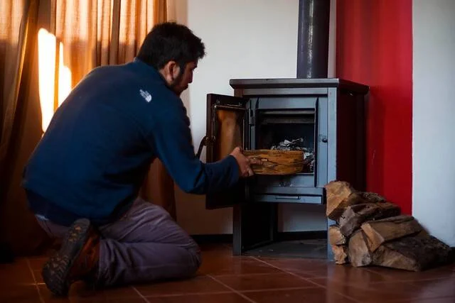 Un hombre arrodillado introduce un trozo de leña en una estufa a leña dentro de una habitación.