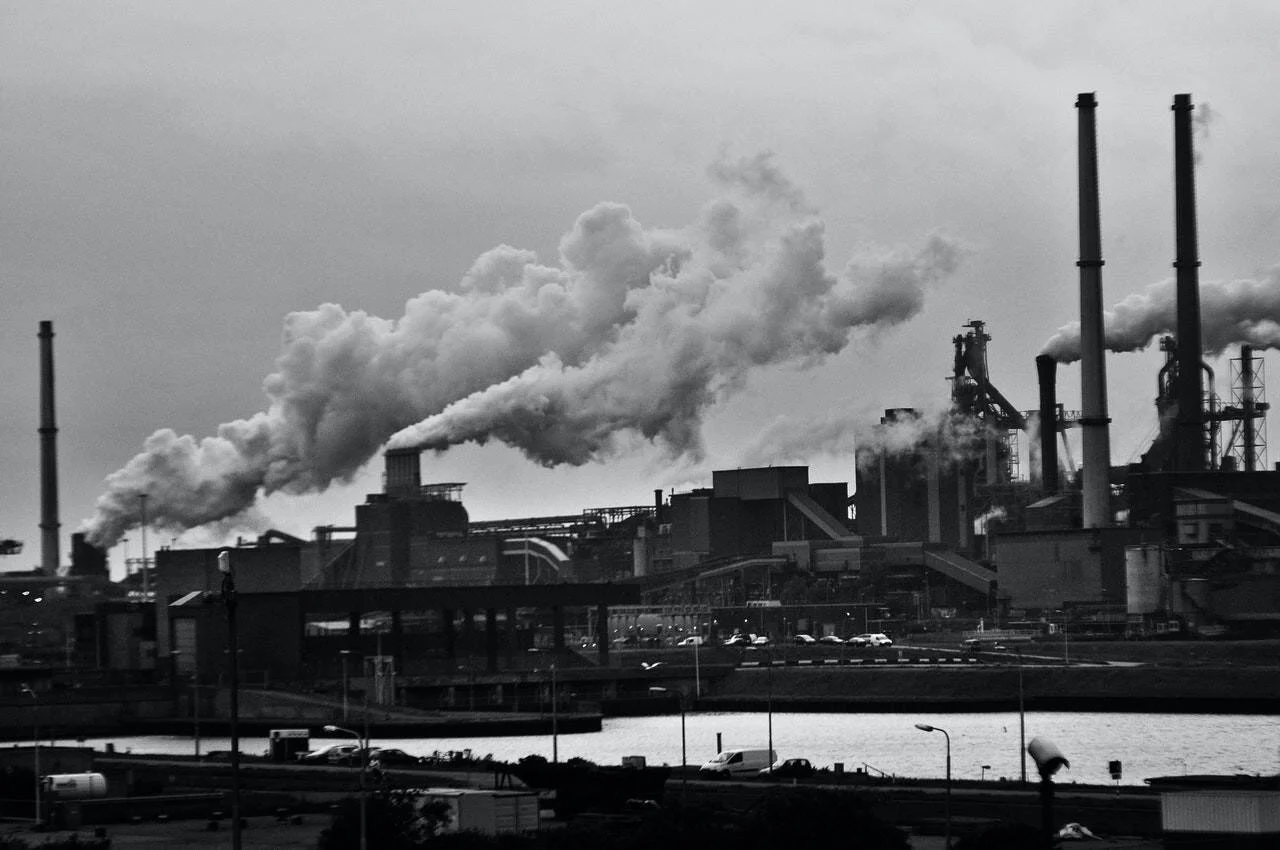 Complejo industrial con múltiples chimeneas expulsando grandes columnas de humo junto a una zona portuaria, en blanco y negro.