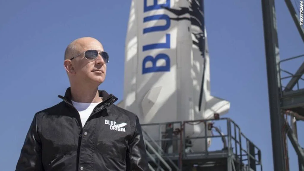 Hombre con lentes de sol y chaqueta de Blue Origin posando frente al cohete New Shepard en la plataforma de lanzamiento.