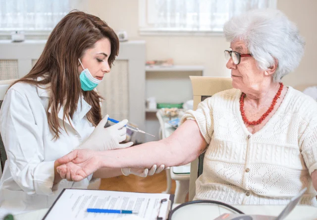Profesional de salud con mascarilla y guantes extrae sangre del brazo de una mujer mayor con gafas.