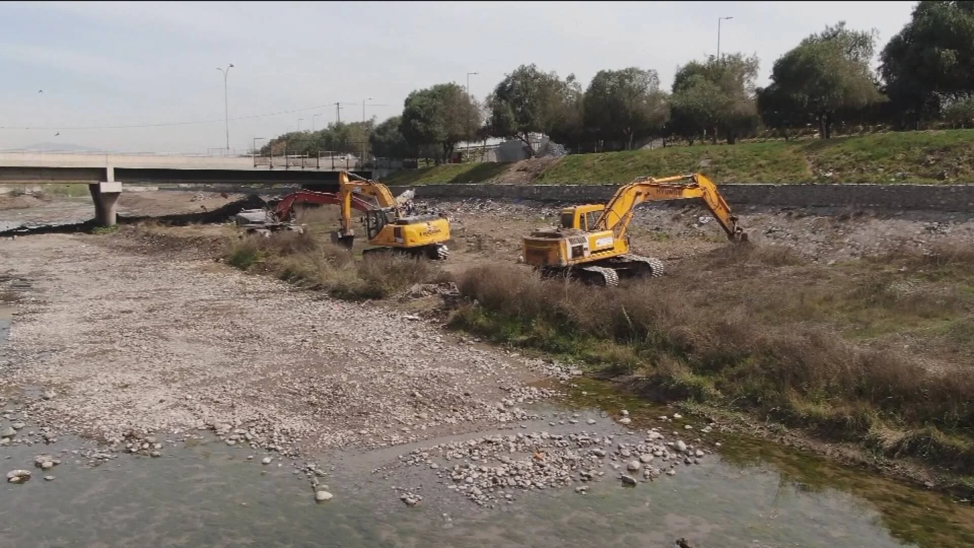 Dos excavadoras amarillas realizan obras de dragado o limpieza en el lecho pedregoso de un río junto a un puente.