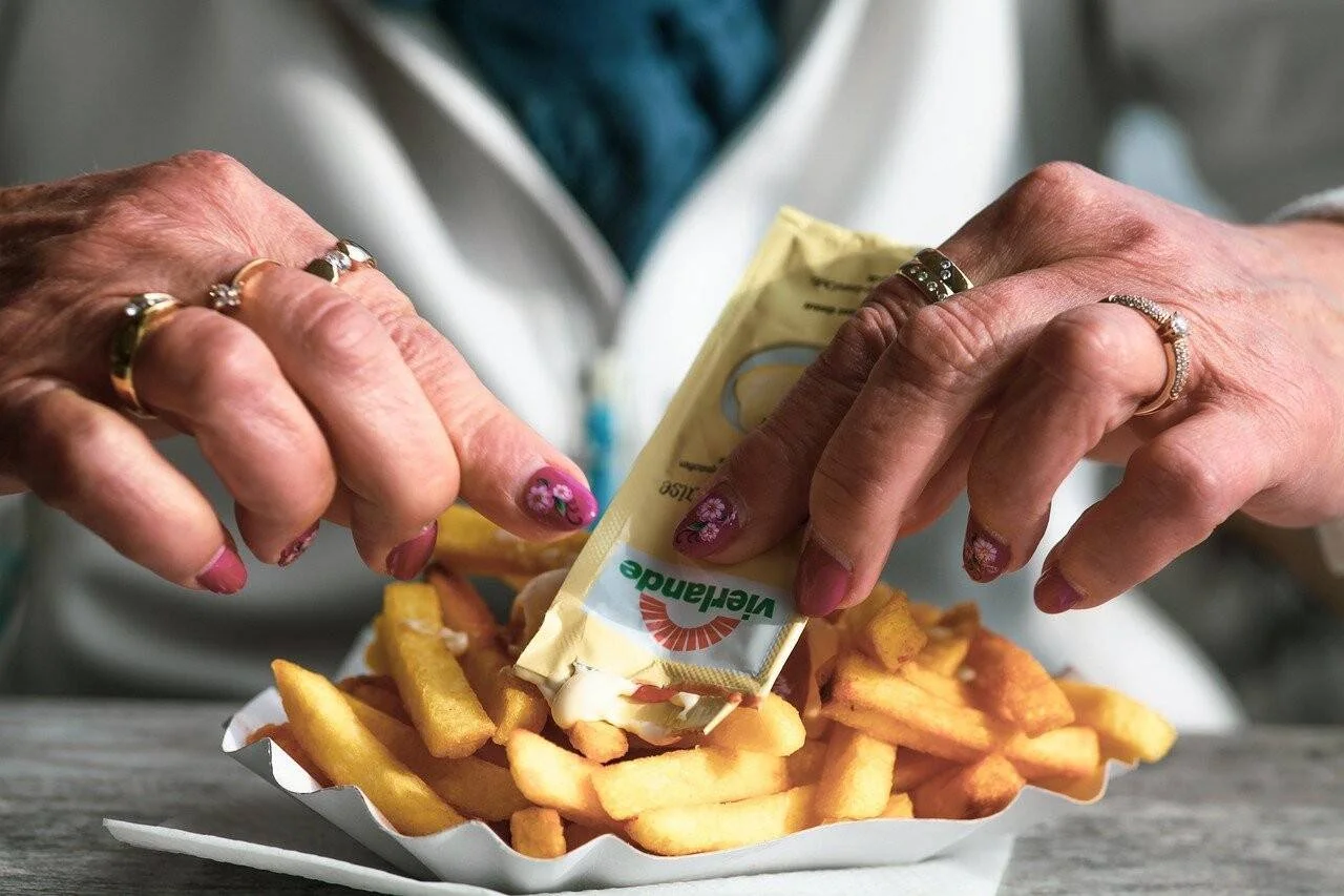 Manos de una persona mayor con anillos abren un sobre de mayonesa sobre una porción de papas fritas en bandeja de cartón.