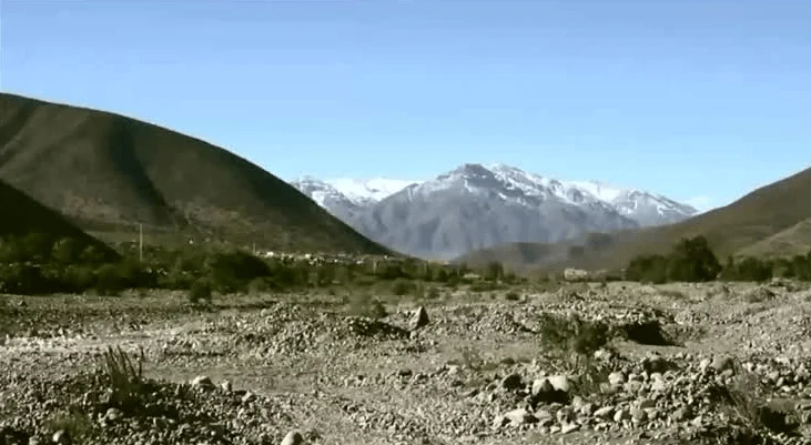 Valle seco y pedregoso rodeado de cerros con cumbres nevadas bajo cielo azul despejado.