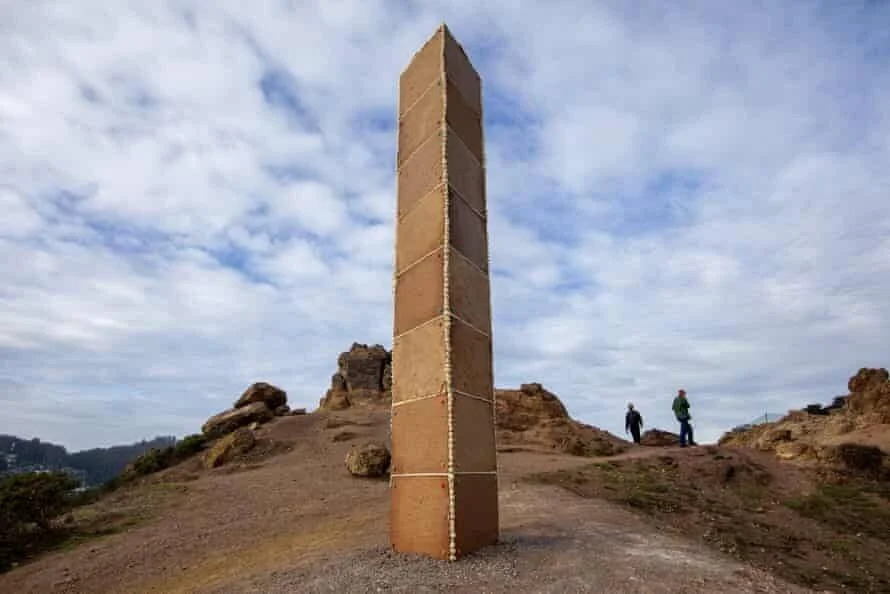 Monolito rectangular de madera apilada y atada con cuerda se alza en una colina rocosa mientras dos personas lo observan al fondo.