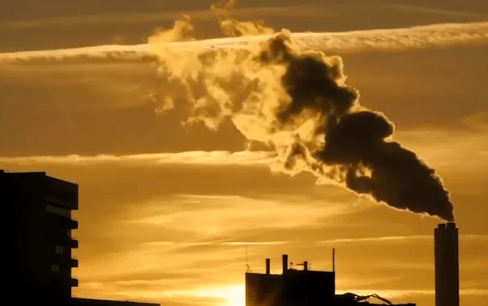 Chimenea industrial expulsa una densa columna de humo frente a un cielo anaranjado al atardecer, con edificios en silueta.