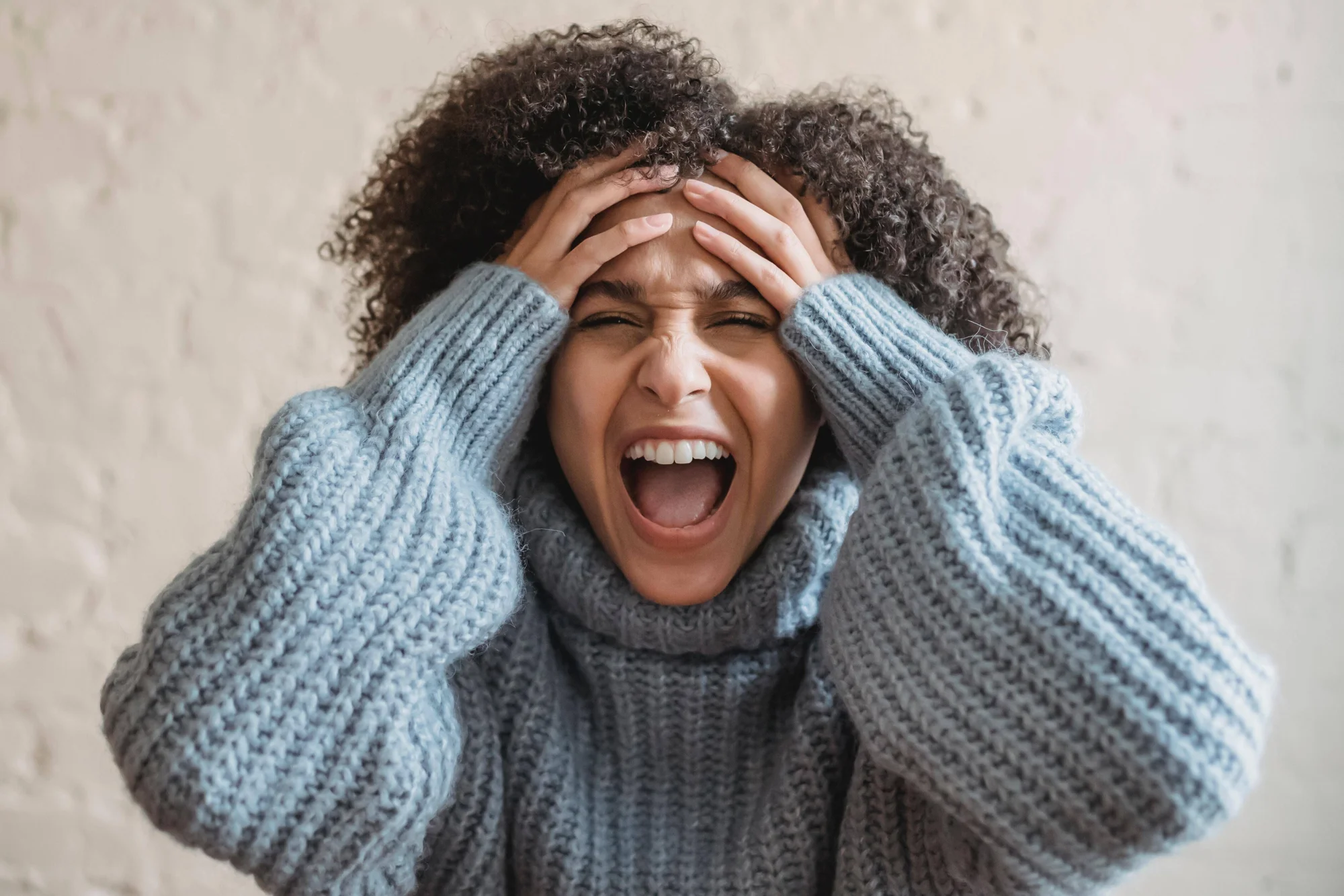 Mujer con suéter gris y cabello rizado grita con los ojos cerrados y las manos apoyadas en la frente, expresando estrés o desesperación.