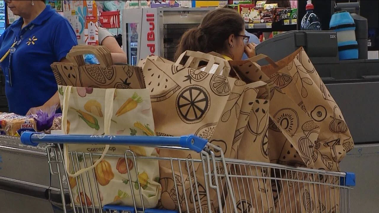 Clienta en caja de supermercado con un carro repleto de bolsas reutilizables de tela y papel, atendida por cajera uniformada.