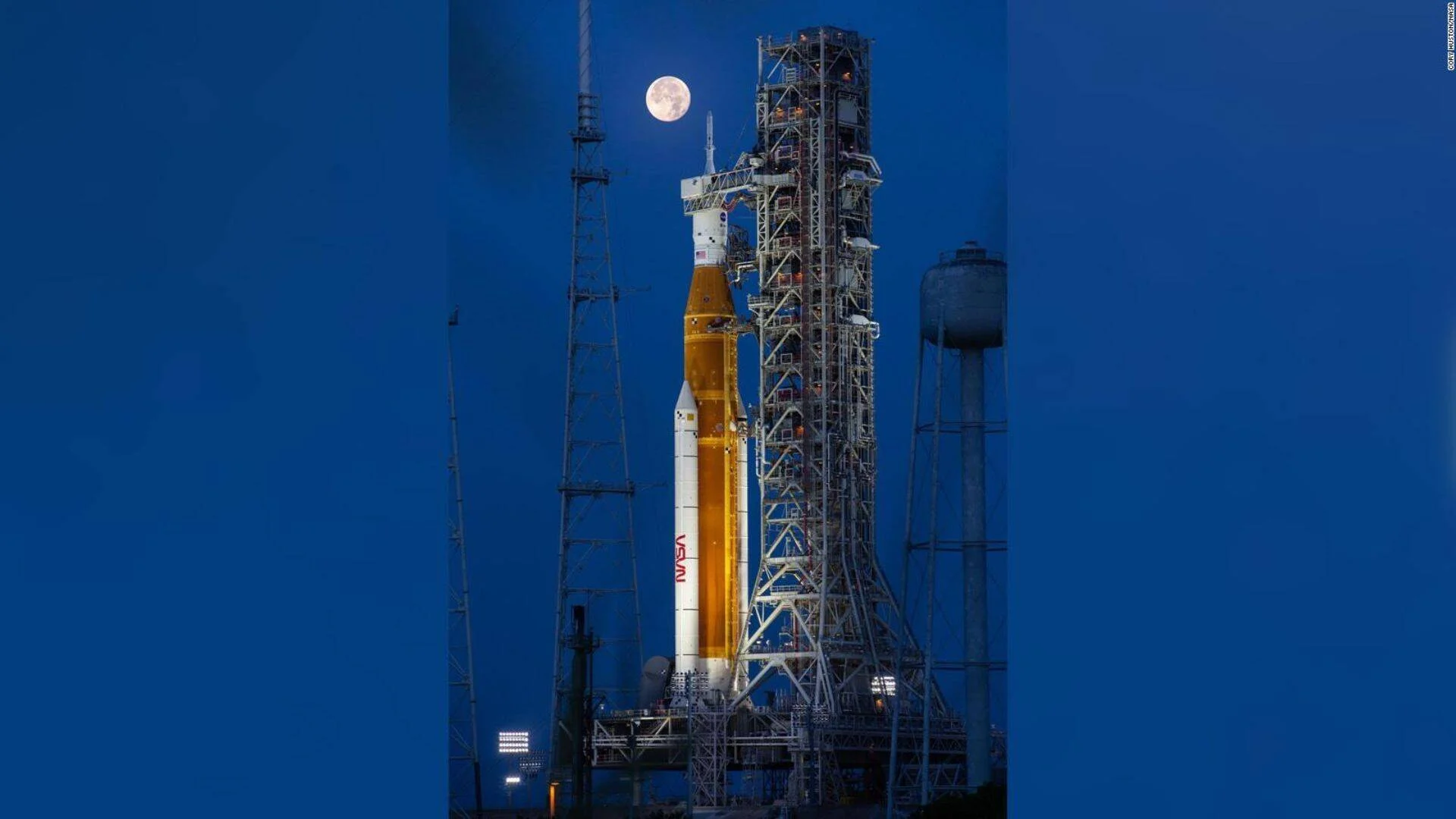 Cohete de la NASA en su plataforma de lanzamiento iluminado de noche, con la luna llena visible al fondo en el cielo azul.