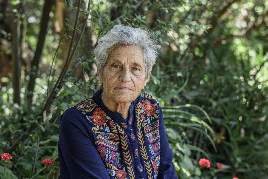Mujer anciana de cabello blanco con blusa azul bordada con flores, posando serena entre vegetación verde.