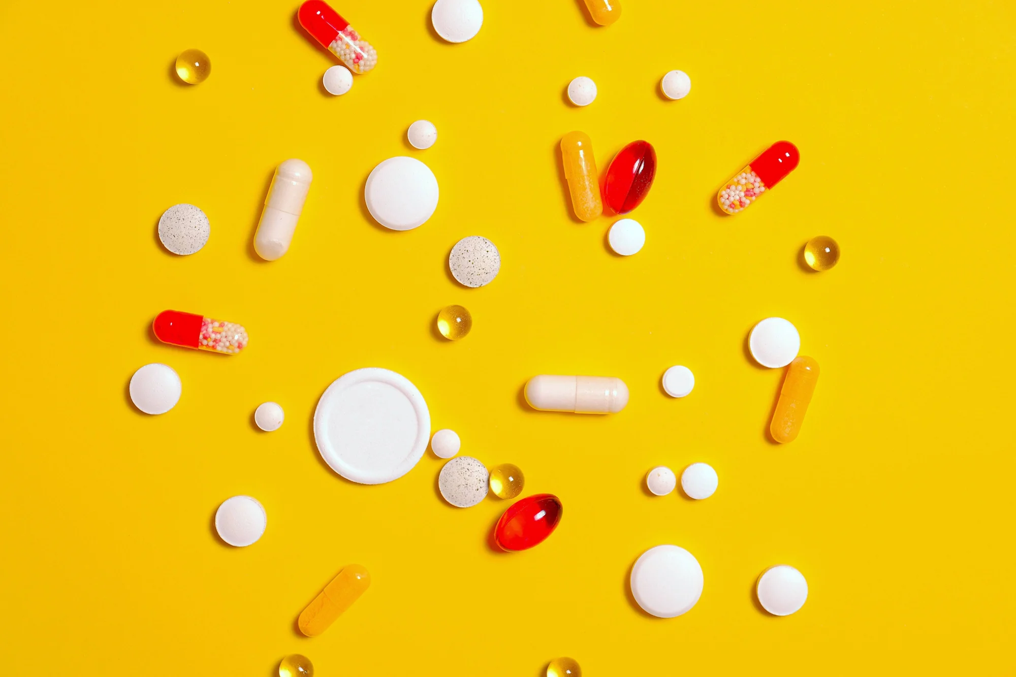 Pastillas, cápsulas y comprimidos de distintos colores y tamaños dispersos sobre una superficie amarilla.
