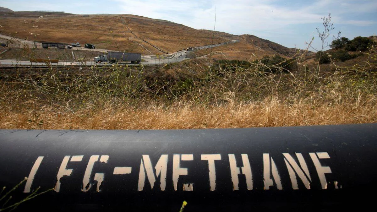 Tubería negra etiquetada 'LFG-Methane' para captura de metano de vertedero, con colinas secas y una autopista al fondo.