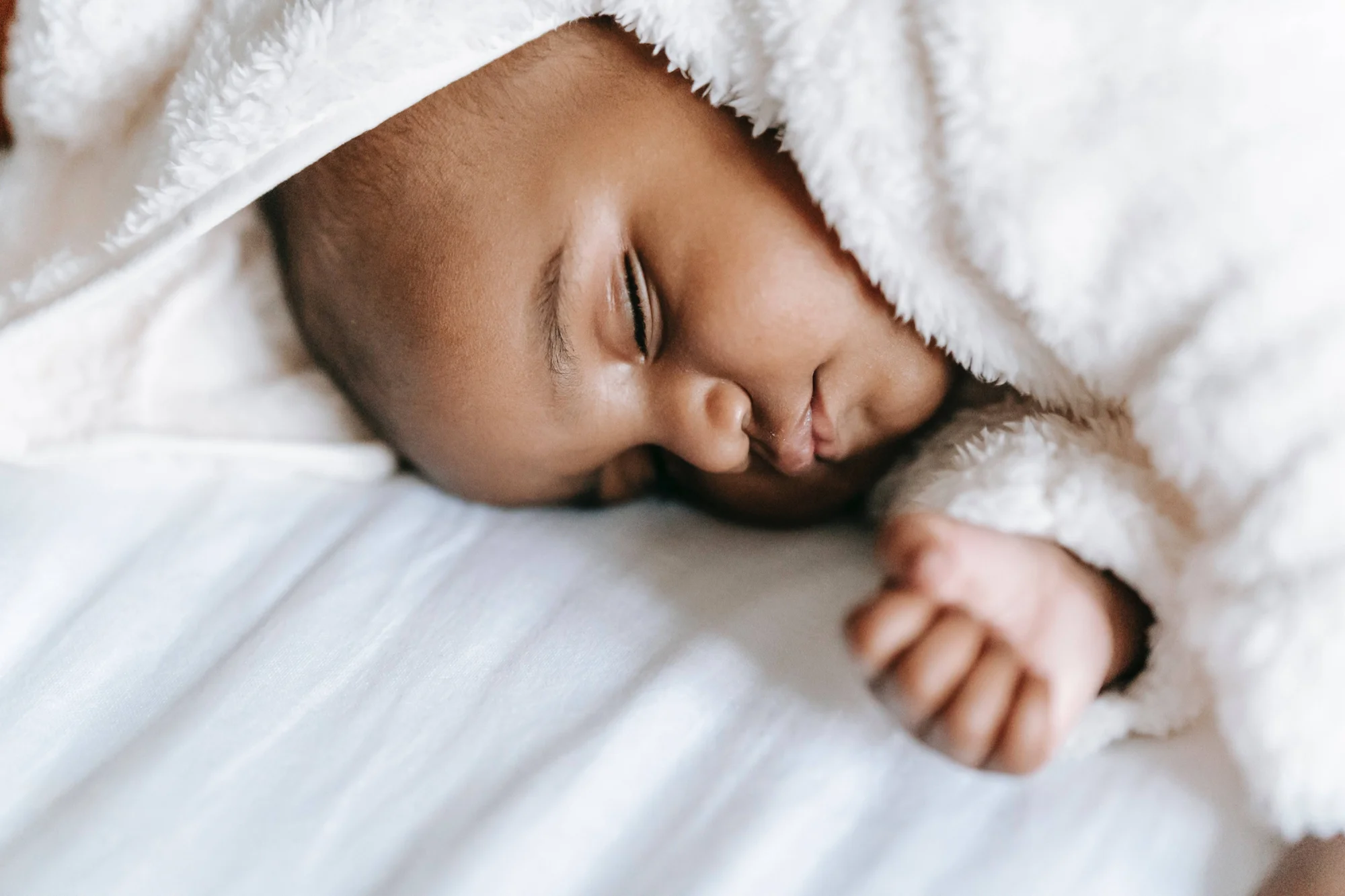 Bebé recién nacido duerme apaciblemente cubierto con una manta blanca esponjosa, con su pequeña mano junto al rostro.