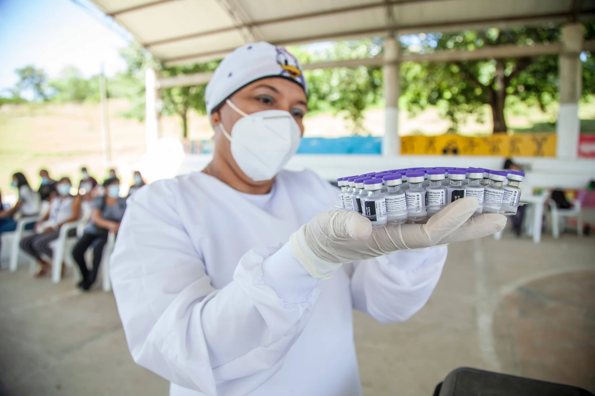 Trabajadora sanitaria con mascarilla y guantes sostiene una bandeja con múltiples viales de vacuna contra el COVID-19 en un puesto de vacunación al aire libre.