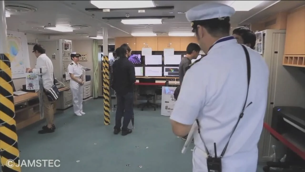 Oficiales y técnicos trabajan en la sala de control de un buque de investigación de JAMSTEC, frente a múltiples monitores.