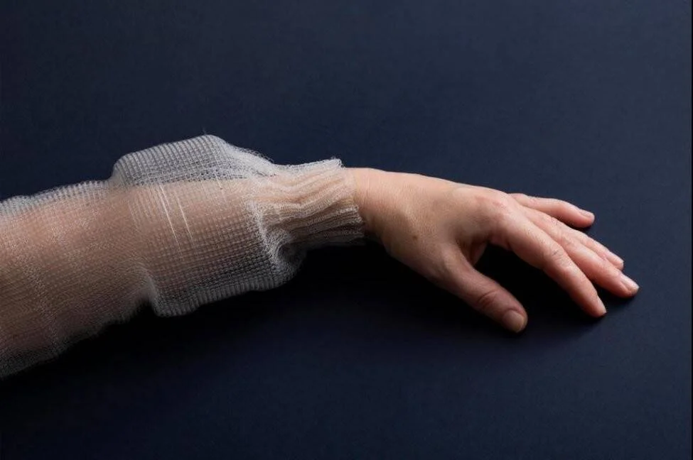Antebrazo y mano de una persona cubiertos con vendas de gasa translúcida sobre fondo azul oscuro.