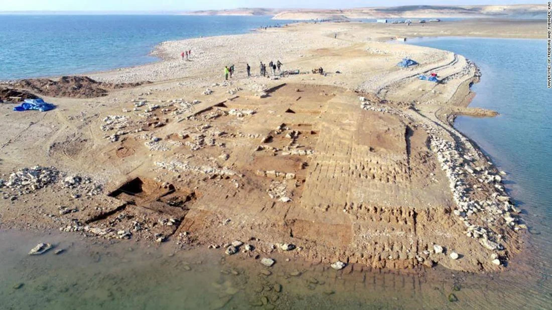 Vista aérea de ruinas de una ciudad antigua expuestas en una península rodeada de agua, con arqueólogos trabajando en el sitio.