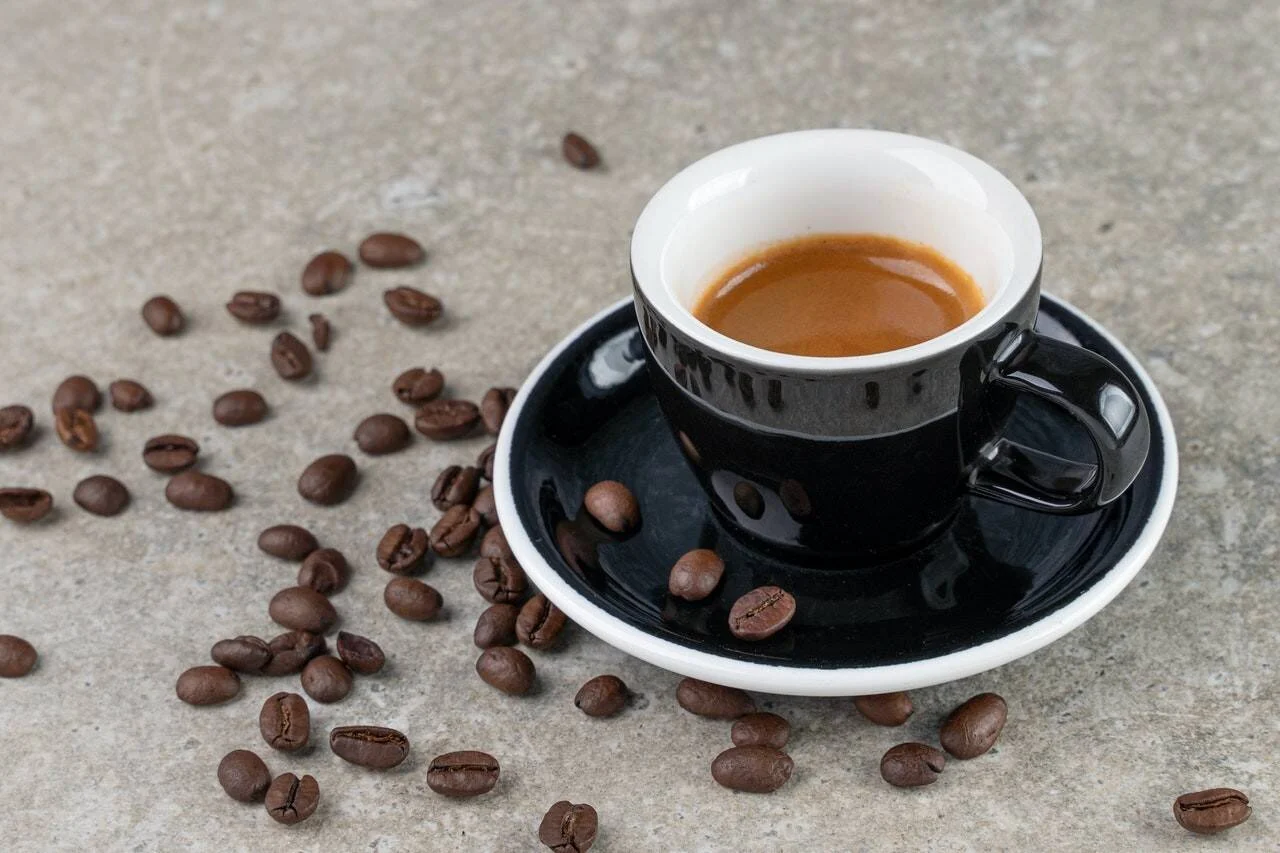 Taza negra de espresso con crema dorada sobre platillo, rodeada de granos de café tostado sobre superficie gris.
