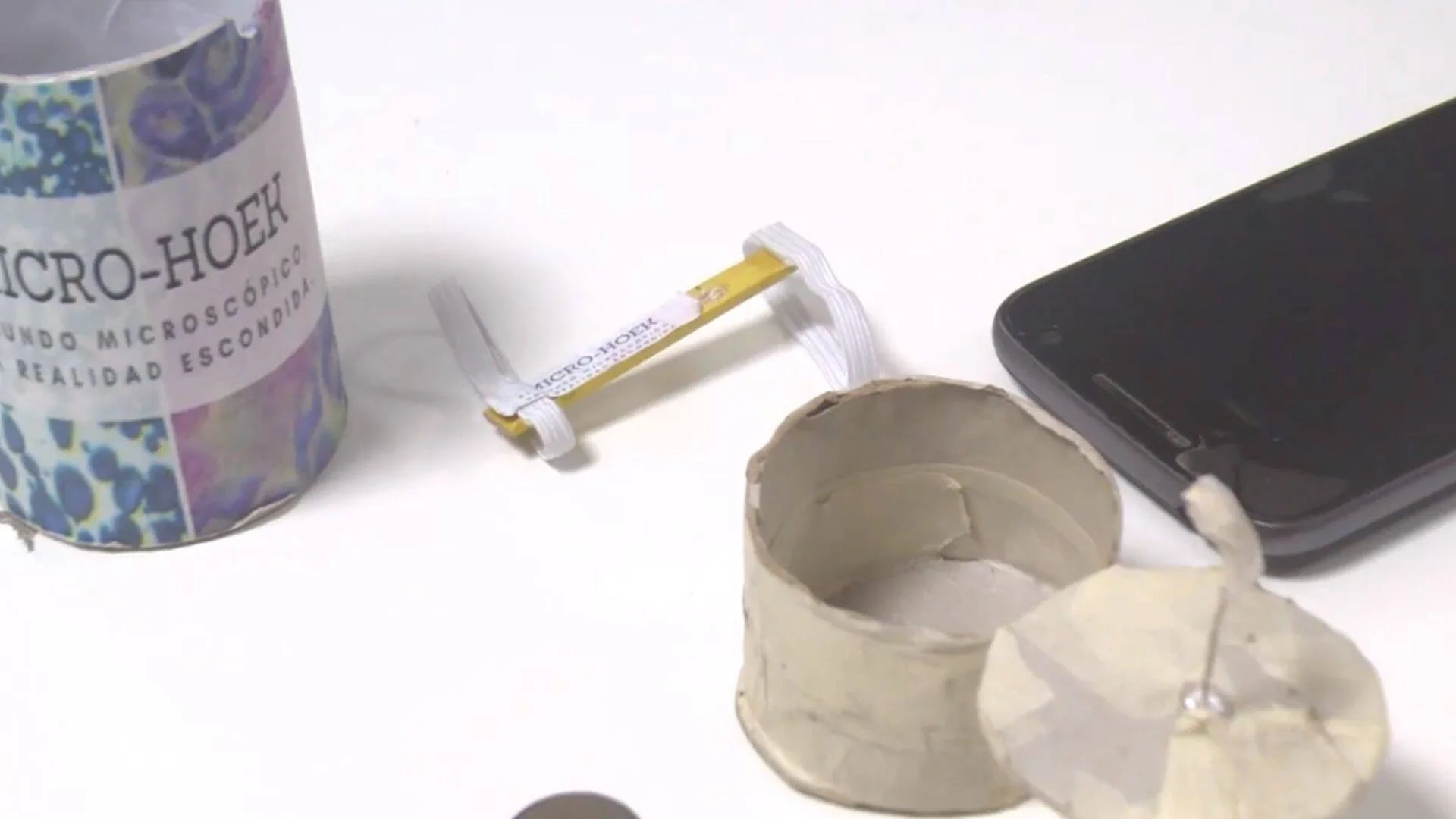 Componentes del kit Micro-Hoek: caja decorada, lente pequeña, soporte de cartón y smartphone sobre superficie blanca.