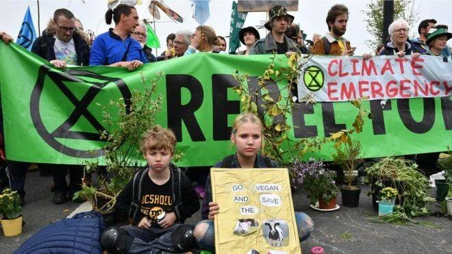 Dos niños sentados frente a una pancarta verde de Extinction Rebellion que dice 'Climate Emergency', sosteniendo un cartel que promueve el veganismo.